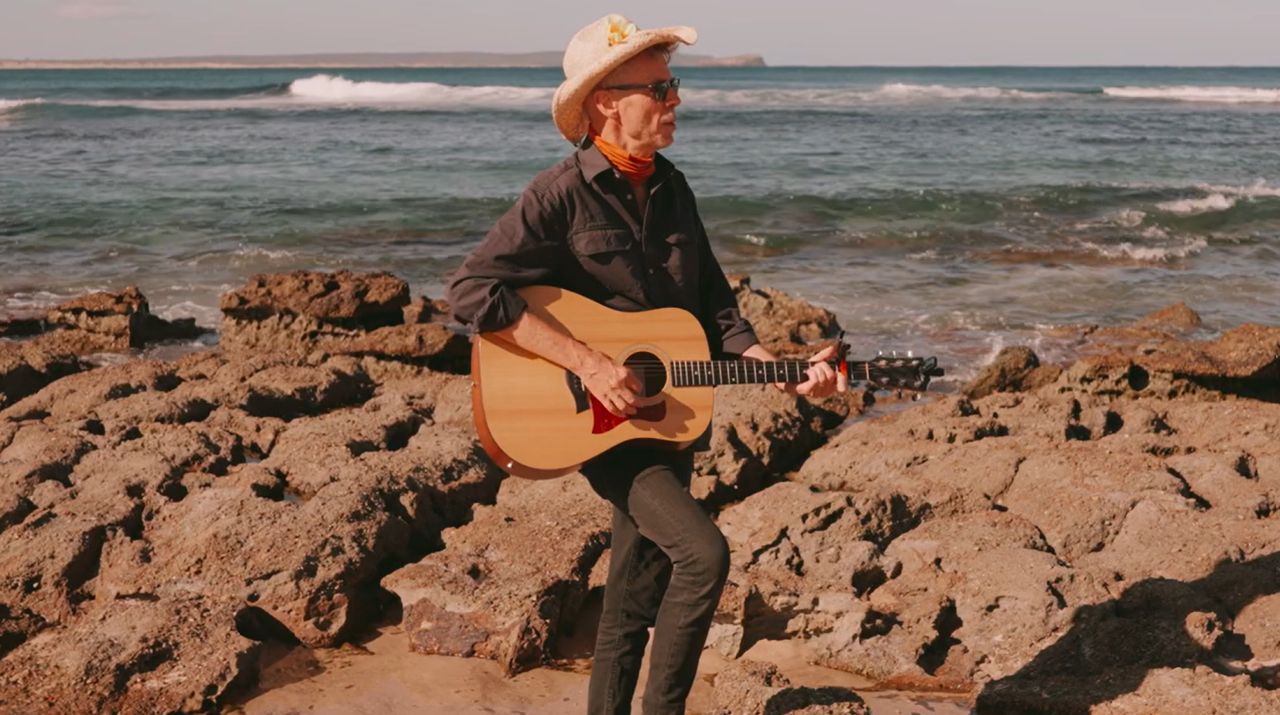 Ian playing acoustic guitar on the rocks by the ocean, from his music video